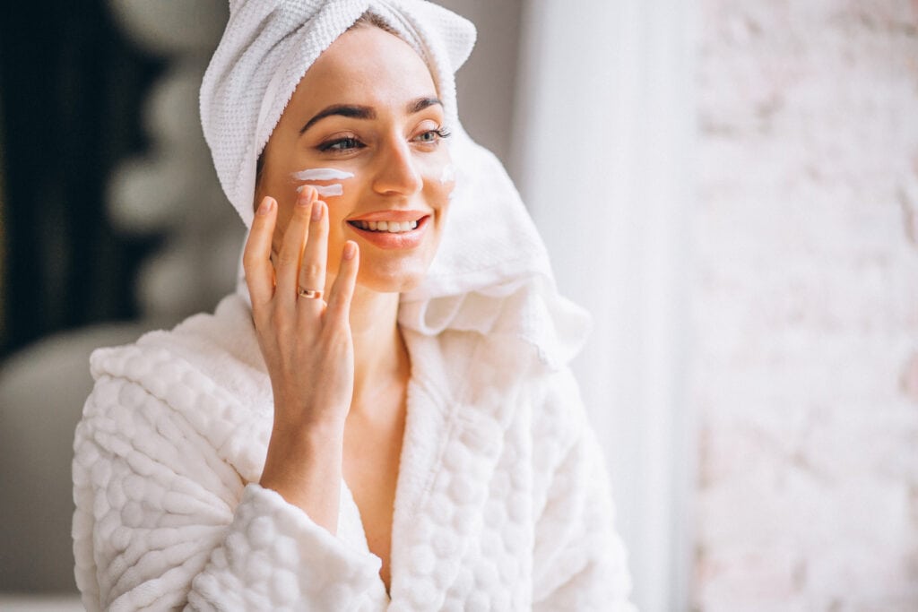 Devojka pre ogledalom nanosi kremu na lice - Make-up saveti za suvu kožu: Kako postići hidrirani izgled zimi