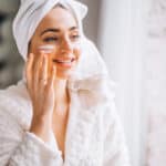 Devojka pre ogledalom nanosi kremu na lice - Make-up saveti za suvu kožu: Kako postići hidrirani izgled zimi